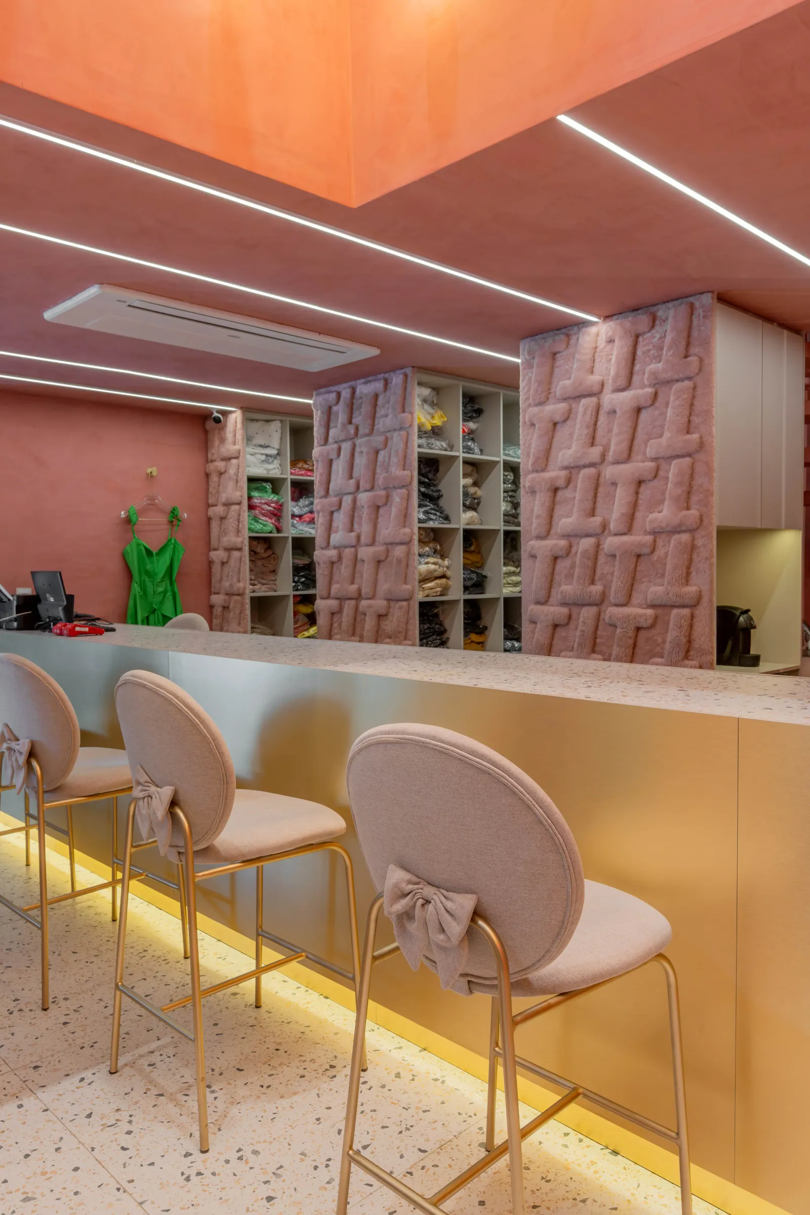 Balcão de atendimento da loja Teca em terrazzo e latão com banquetas rosa de laço, nichos organizando peças dobradas embaladas, painéis de pelúcia rosa com monograma T e vestido verde pendurado