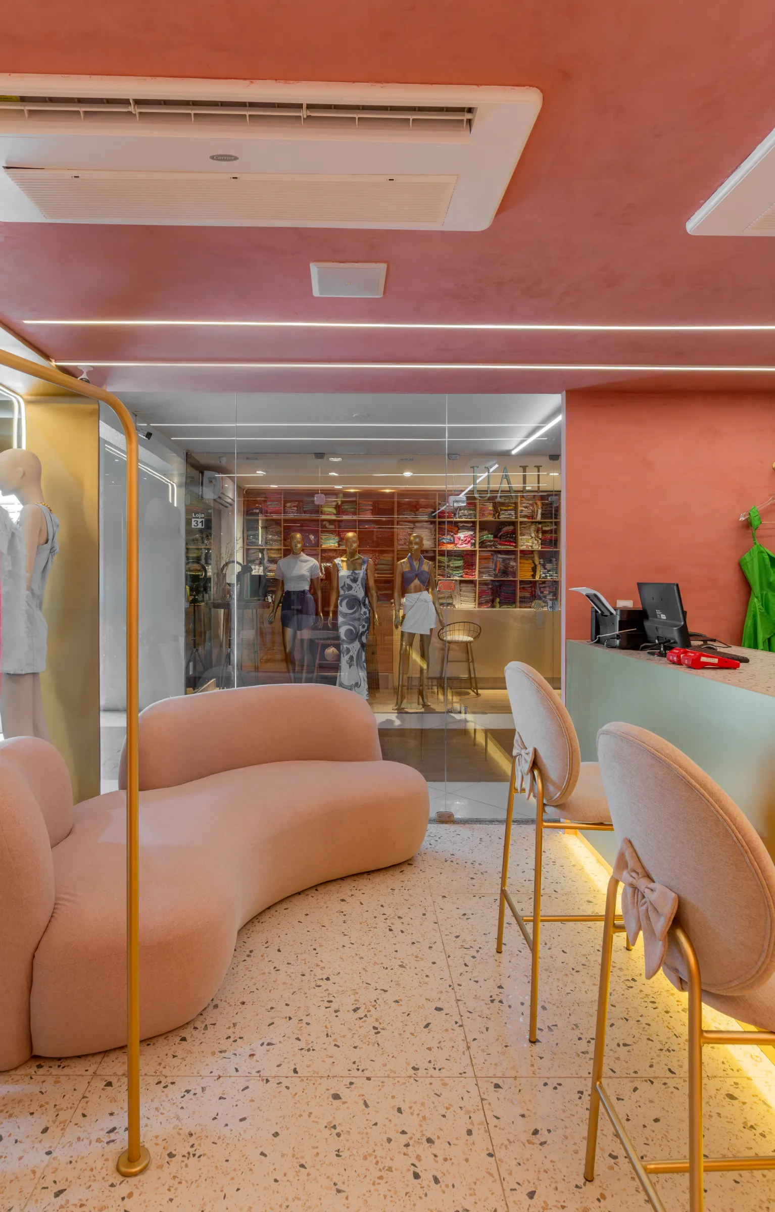 Ambiente da loja Teca com sofá curvo rosa, banquetas de laço, teto terracota e vitrine de vidro com manequins vestidos e o logotipo da marca ao fundo