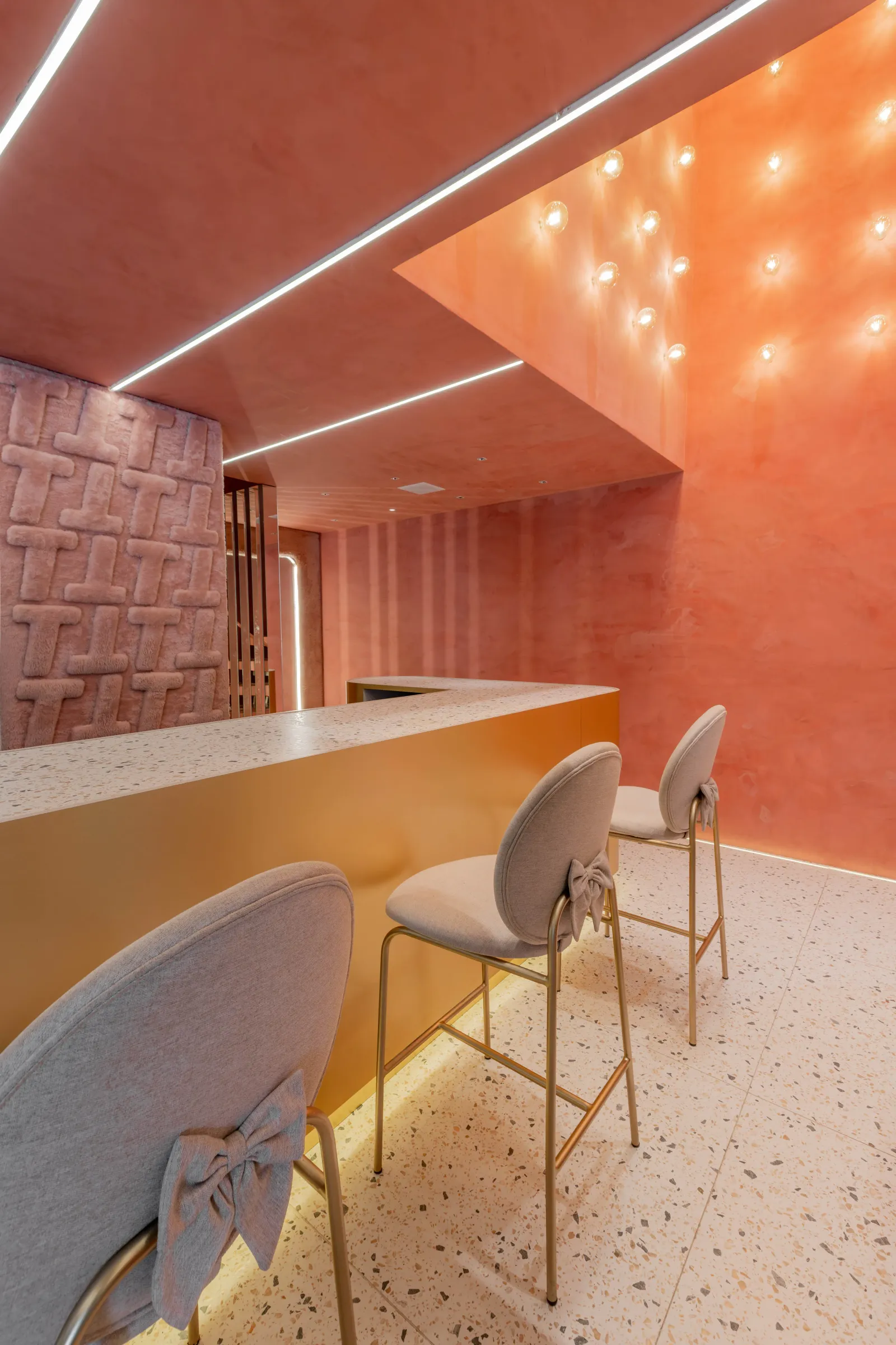 Vista do balcão de terrazzo e latão da loja Teca com banquetas rosa de laço, teto terracota com recortes geométricos e lâmpadas acesas e painel de pelúcia rosa com monograma T à esquerda
