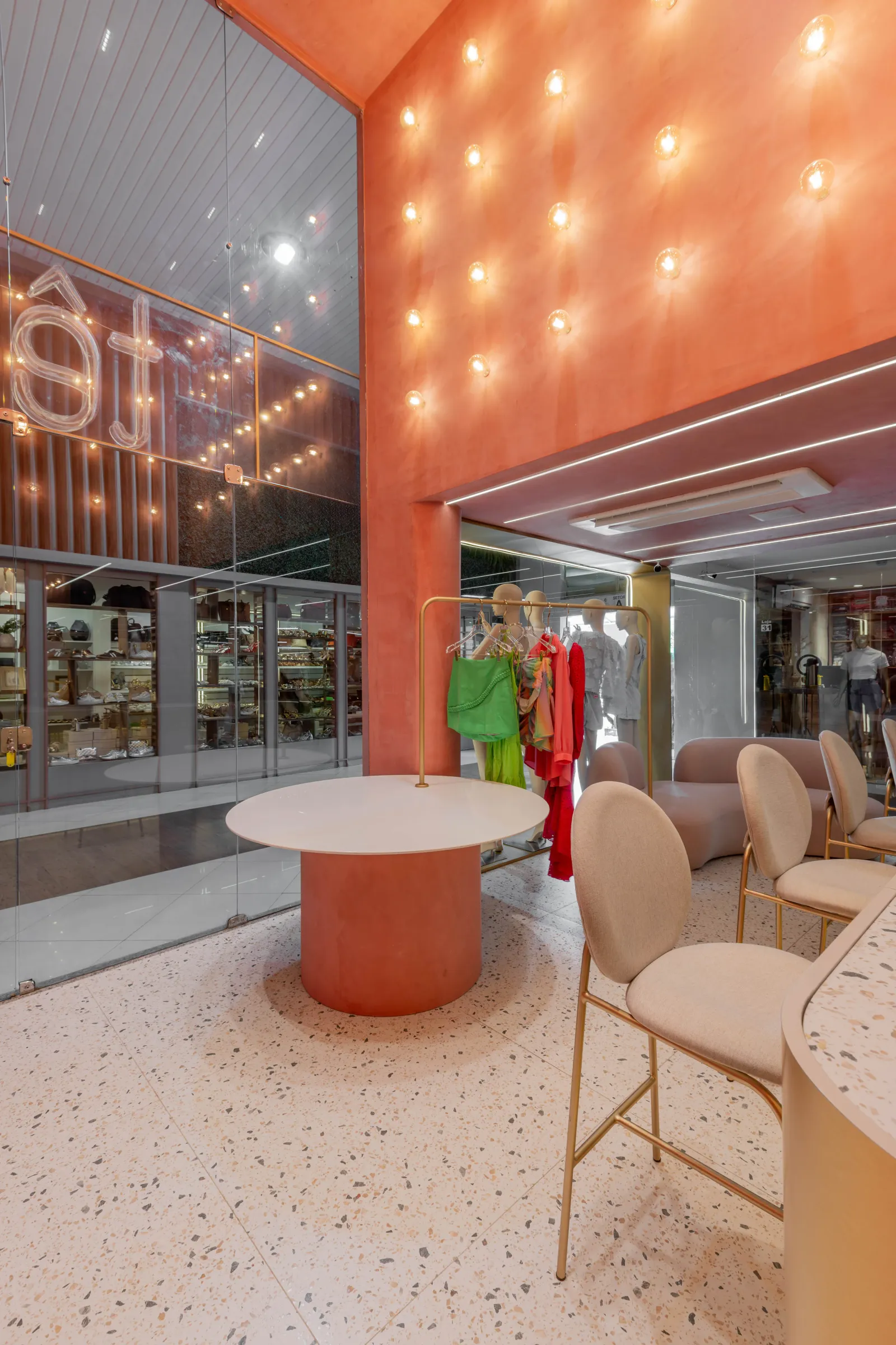 Detalhe de puxador oval em latão com miolo de terrazzo numa porta de vidro da loja Teca, com corredor terracota e nicho iluminado ao fundo